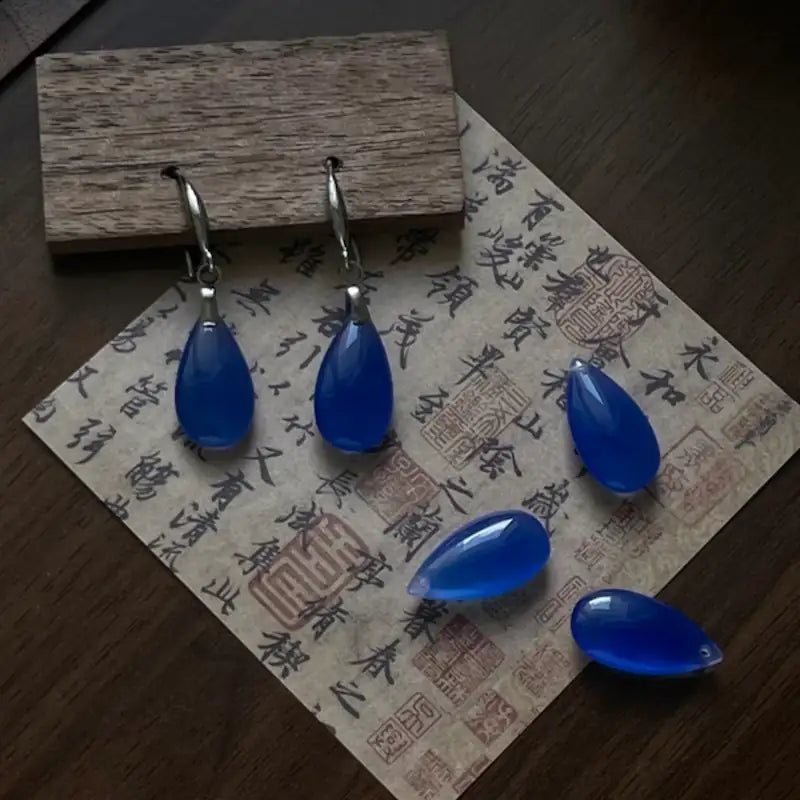 Orienlu flat lay display of silver-plated hook teardrop earrings alongside loose blue agate gemstone beads on vintage parchment paper, emphasizing the authentic mineral clarity and color consistency.