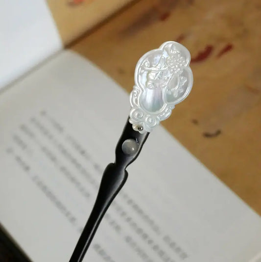 Macro close-up of the Orienlu wooden hair pin head resting over an open book, showcasing the iridescent pearlescent luster of the carved shell inlay and the smooth grain of the black ebony wood.