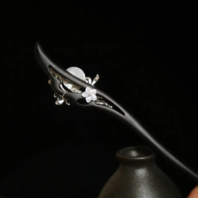 The reverse side view of the Orienlu hair pin inserted in a ceramic vase, revealing a delicate carved white shell flower embedded seamlessly within the hollow ebony frame.