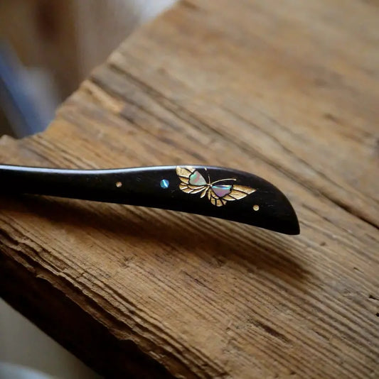 A macro close-up detail shot of the Orienlu ebony hair stick, focusing on the shimmering pink and green iridescence of the inlaid shell butterfly charm resting on a rustic wooden surface.