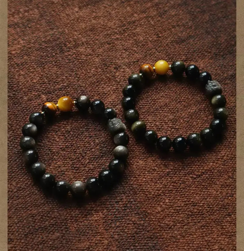 Two Orienlu silver sheen obsidian beaded bracelets laid side-by-side on brown canvas, highlighting the brilliant chatoyancy of yellow tiger's eye and the metallic alloy beast spacers.