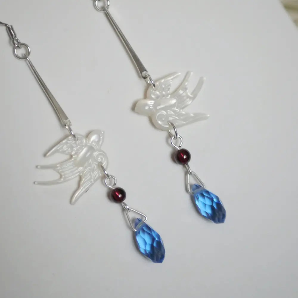 Close-up of carved mother of pearl white swallow, red garnet bead, and faceted blue glass teardrop on long drop earrings — Orienlu