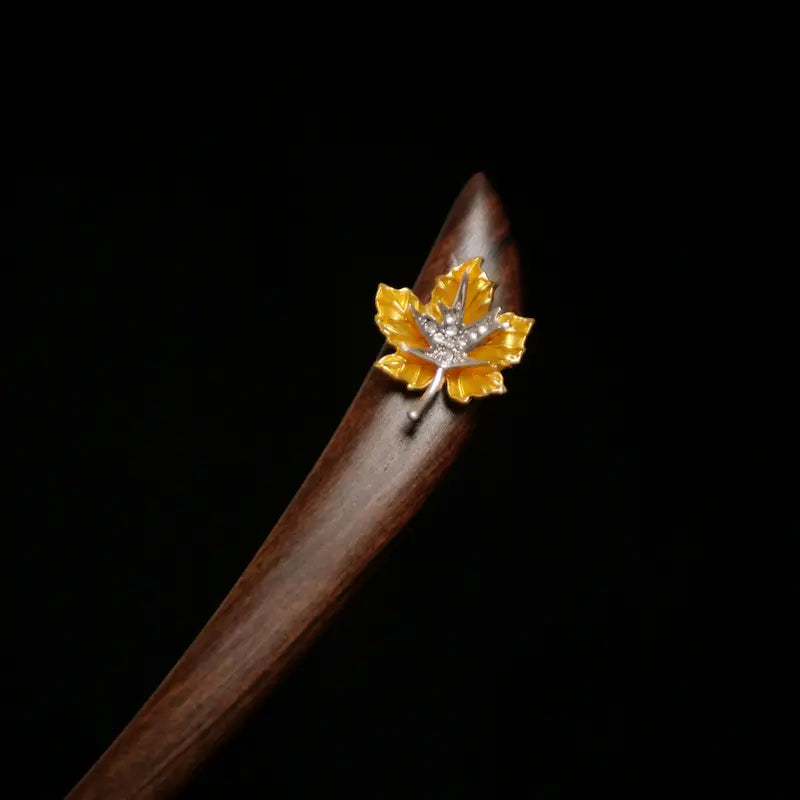 An extreme close-up shot focusing on the exquisite craftsmanship of the copper enamel maple leaf on the dark timber hair stick, showing the vibrant golden glaze and crystal pave veins.