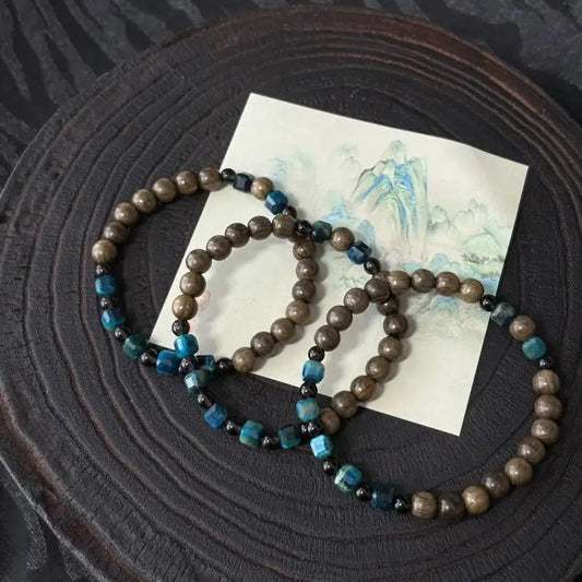 Three Orienlu wooden meditation bracelets stacked on a vintage painting card, highlighting the chatoyancy of blue-green hawk's eye stone cubes and glossy smoky quartz spacer beads.