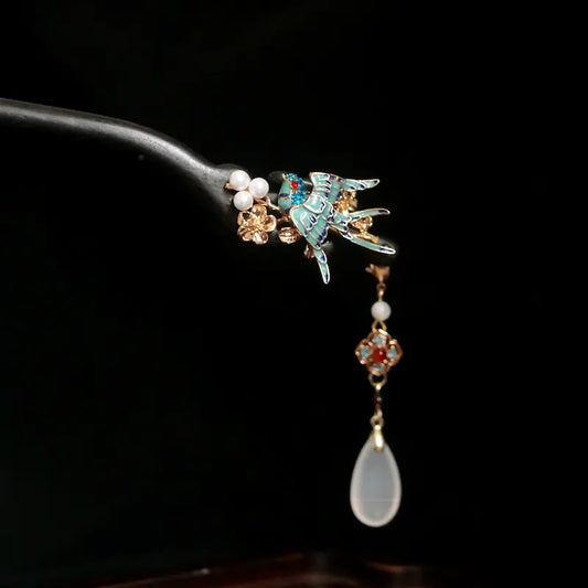 A macro close-up detail shot of the Orienlu sandalwood hair stick, showcasing the vibrant enamel craftsmanship of the cloisonne bird, cluster of white shell pearls, and a hanging tassel with a translucent teardrop agate pendant against a black background.