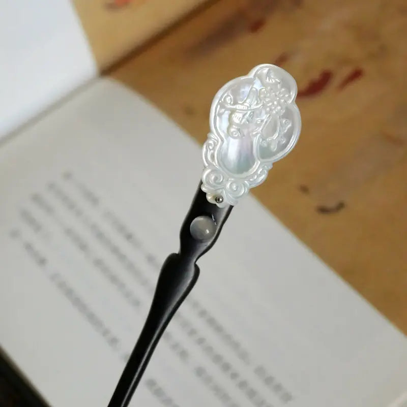 Macro close-up of the Orienlu wooden hair pin head resting over an open book, showcasing the iridescent pearlescent luster of the carved shell inlay and the smooth grain of the black ebony wood.