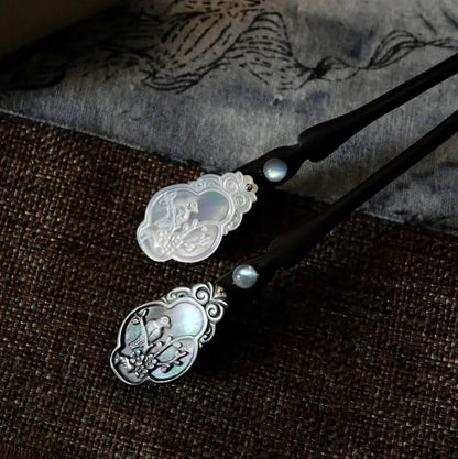 Two Orienlu natural ebony hair sticks laid flat on a vintage Asian art print background, emphasizing the unique labradorescence of the flash gemstone beads and the intricate magpie craftsmanship.