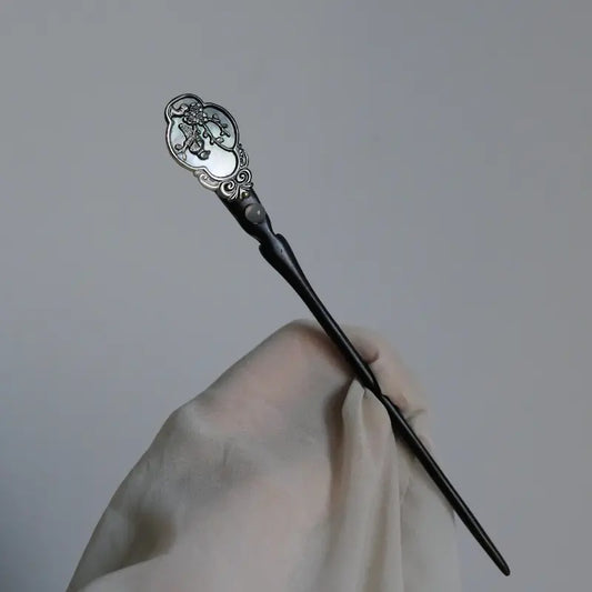 Orienlu product shot of a hand holding an ebony wood hair stick against a light gray background, showing the complete sleek silhouette, a carved mother-of-pearl ruyi top, and an inset cabochon labradorite stone.