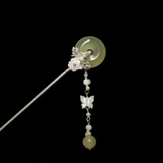 Orienlu product shot of a silver-toned hair stick against a solid black background, showcasing a translucent light green agate donut disc accented with a dangling mother-of-pearl butterfly tassel.