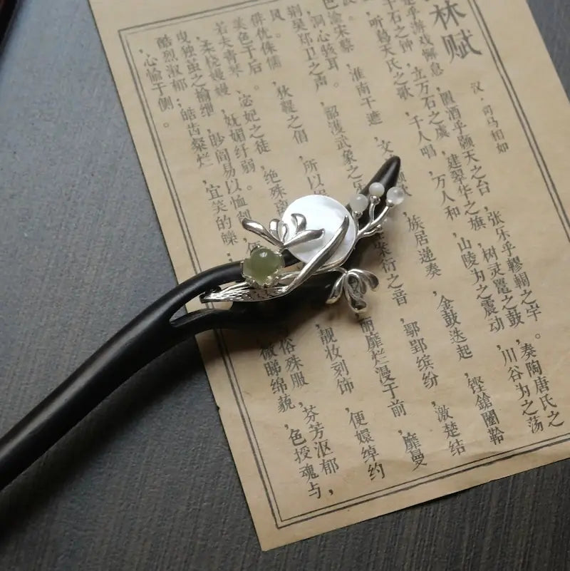 A top-down lifestyle shot of the Orienlu jade and shell wooden hair stick resting on an ancient Chinese calligraphy scroll, showcasing the detailed silver metalwork.