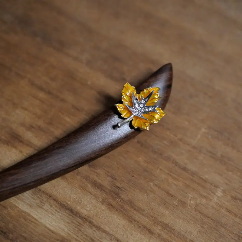 A top-down view of the custom-cut autumn maple leaf wooden hair stick resting on a natural grain tabletop, showcasing the polished dark red tones of premium sandalwood.