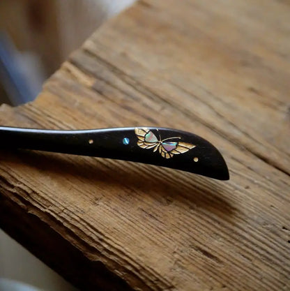 A macro close-up detail shot of the Orienlu ebony hair stick, focusing on the shimmering pink and green iridescence of the inlaid shell butterfly charm resting on a rustic wooden surface.