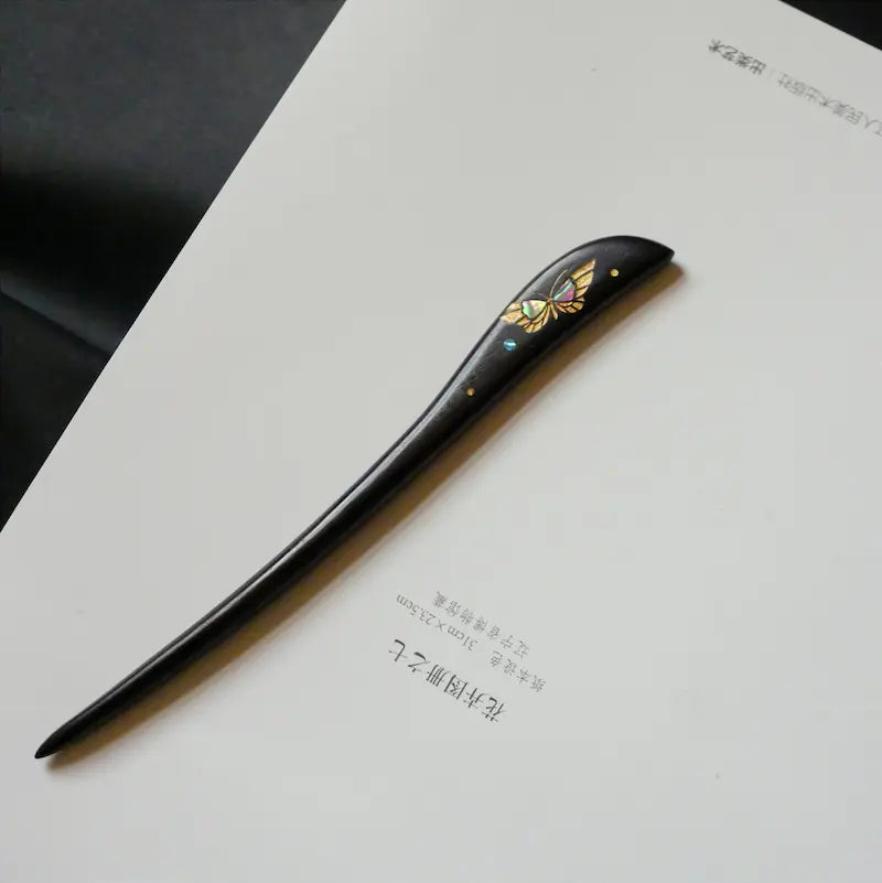 An angled top-view display of the Orienlu raden shell butterfly hair stick resting on a minimalist white paper background, highlighting its sleek contours and organic texture.