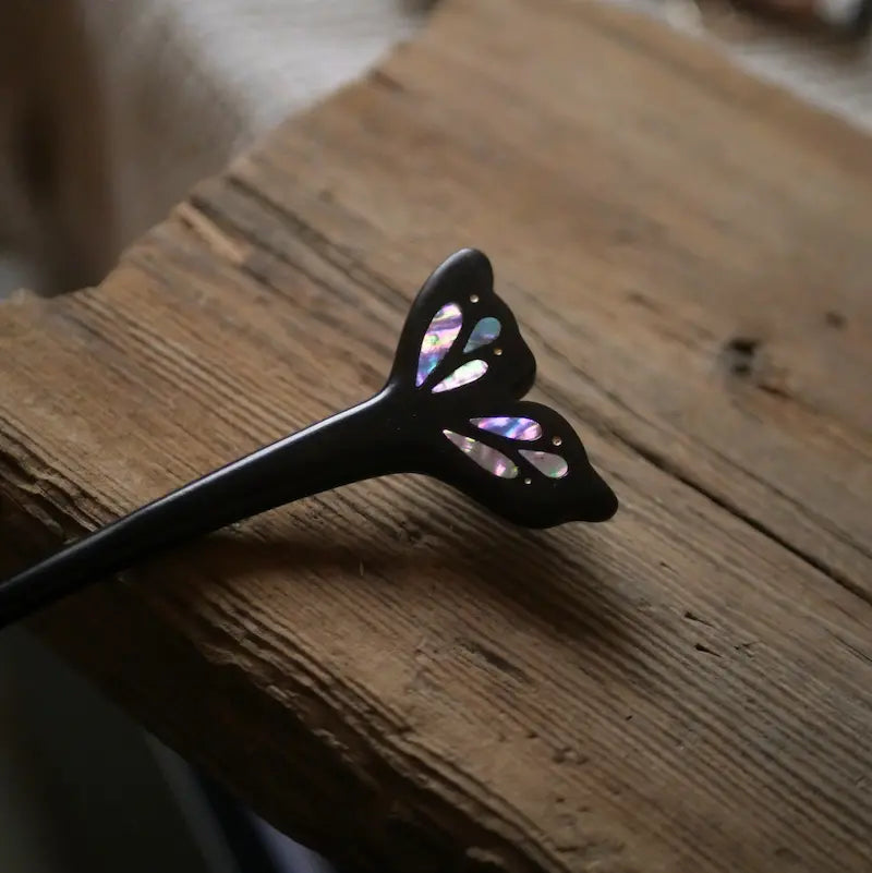 A macro close-up detail shot of the Orienlu ebony hair stick, capturing the shimmering pink and purple iridescence of the raden shell inlay on a rustic wooden surface.