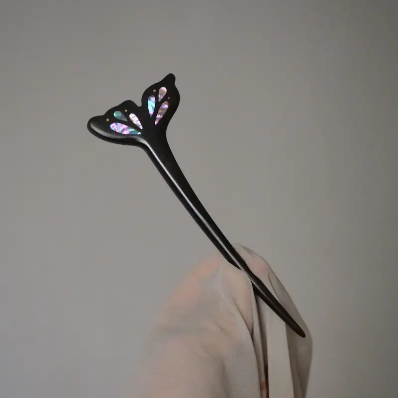 An angled hand-held display of the Orienlu mother of pearl inlaid ebony wood hair jewelry against a neutral grey studio background, highlighting its sleek contours.