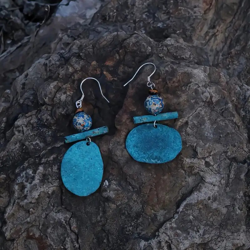 Orienlu alternative product angle of the verdigris bronze and patterned lacquerware dangles resting on a rugged, dark volcanic rock matrix, emphasizing the tactile, weathered crust of the oxidized bronze and the organic curves of the pure silver ear hooks.