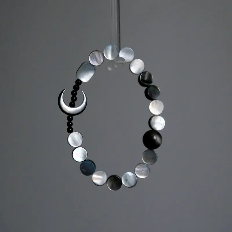 A hanging view of the Orienlu silver sheen obsidian and mother of pearl bracelet, showcasing the shimmering iridescence of the flat round shell beads and the sleek crescent moon against a minimalist grey background.