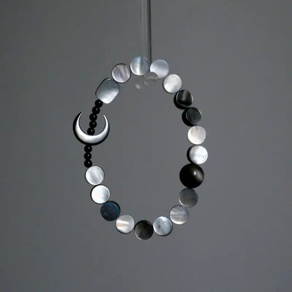 A hanging view of the Orienlu silver sheen obsidian and mother of pearl bracelet, showcasing the shimmering iridescence of the flat round shell beads and the sleek crescent moon against a minimalist grey background.