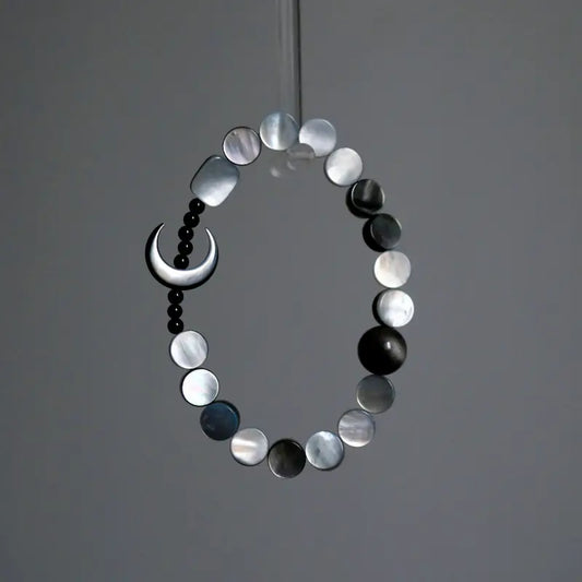 A hanging view of the Orienlu silver sheen obsidian and mother of pearl bracelet, showcasing the shimmering iridescence of the flat round shell beads and the sleek crescent moon against a minimalist grey background.