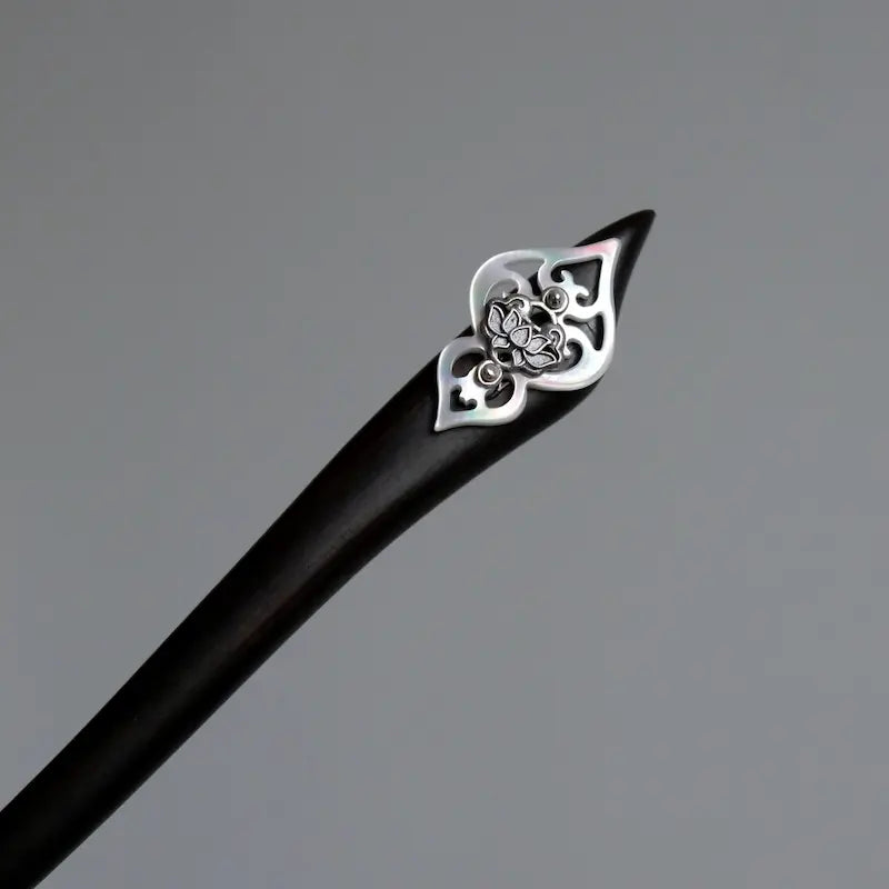 An overhead macro studio shot focusing on the geometric cut of the black timber hair pin topped with solid silver craftsmanship and a carved white shell base.