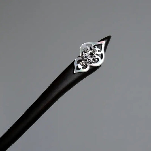 An overhead macro studio shot focusing on the geometric cut of the black timber hair pin topped with solid silver craftsmanship and a carved white shell base.